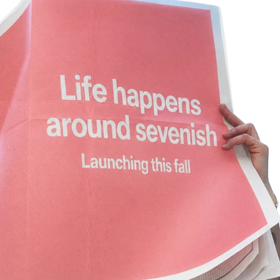 Someone holding up a coral newspaper that reads: Life happens around sevenish. Launching this fall.